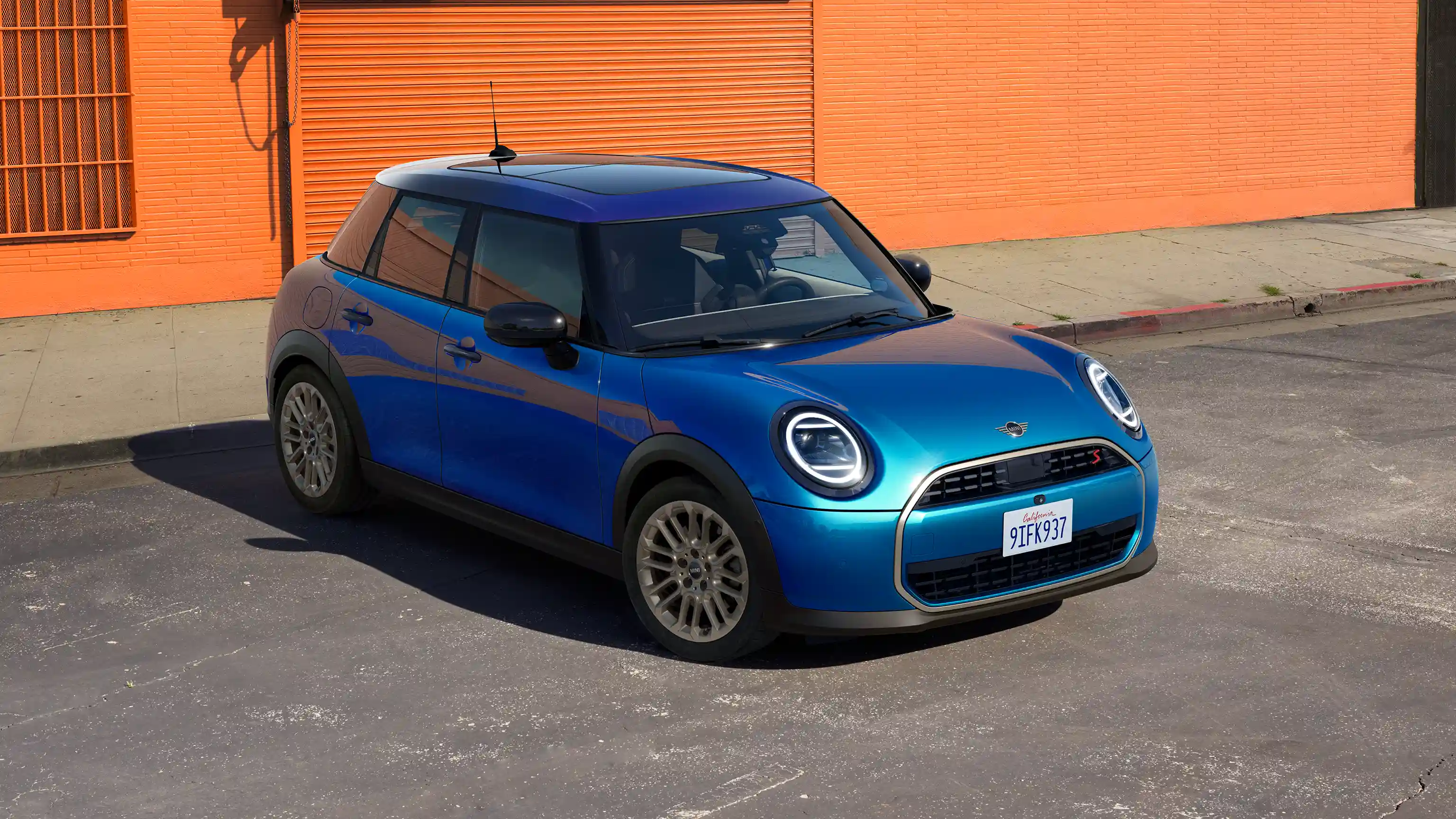 Front view of a MINI Cooper 5-Door in Icy Sunshine Blue and Multitone Blue Roof parked on the street with orange wall in the background