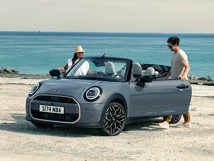 Front left view of the MINI Cooper Convertible in Copper Grey at a beach with two people next to it.