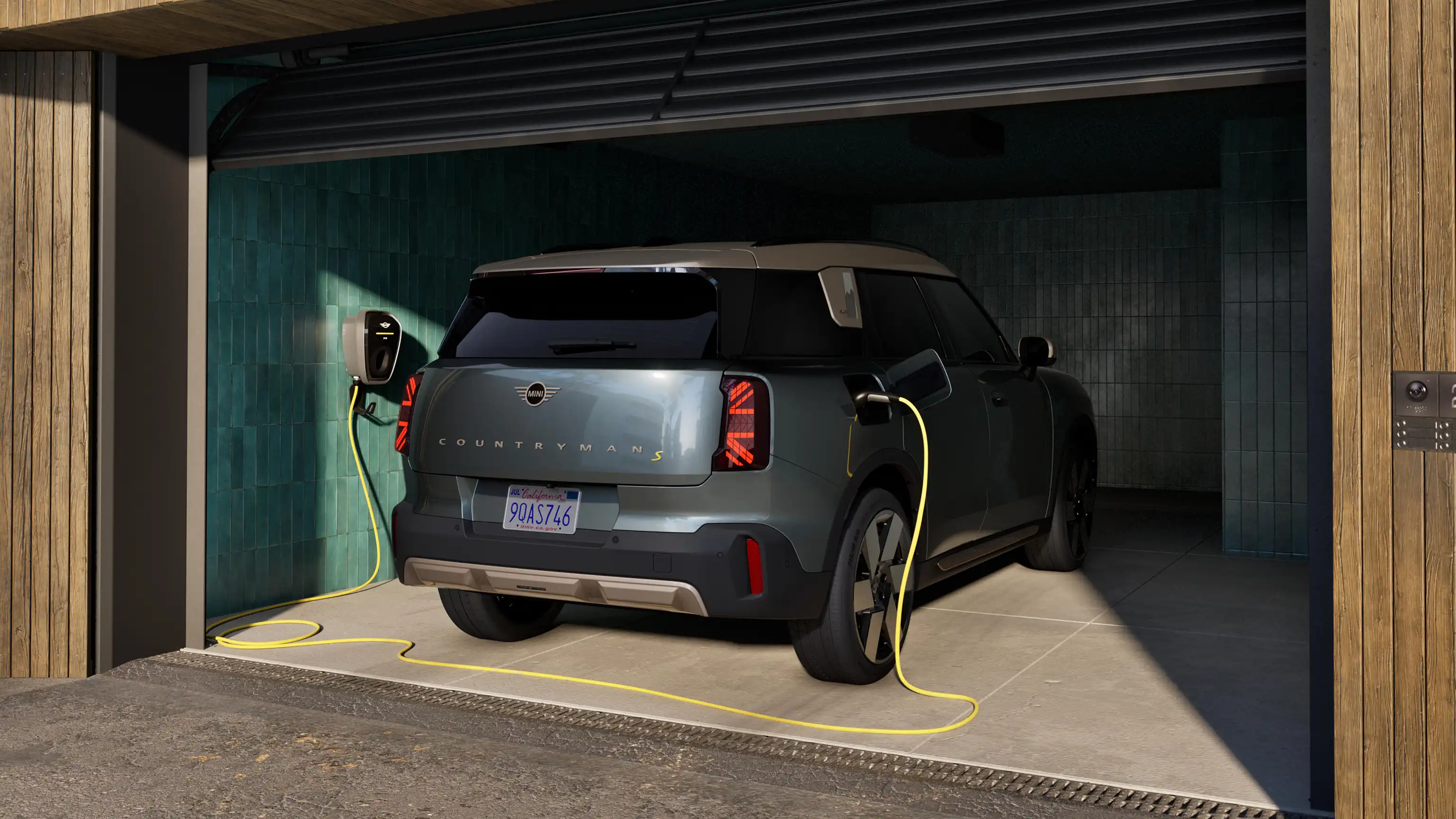 Rear view of a charging all-electric MINI Countryman in Smokey Green standing in open garage connected with charging cable.